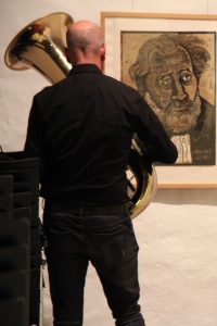 Ein Musiker (Rückenansicht) mit Tuba spielt vor einem Selbstbildnis Otto Pankoks.