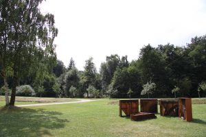 Vier Klaviere stehen in einer sonnigen Parklandschaft (Gras, Bäume), davor liegt ein Flügel.