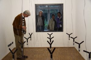 Eine kinetische Klanginstallation, von einem Besucher intensiv betrachtet, im Hintergund schauen drei weitere Besucher durch ein Fenster zu.