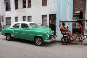Grünweißer Oldtimer (Chevrolet) in Havanna und dreirädriges Fahrradtaxi mit Mutter und zwei Kindern stehen am Straßenrand.