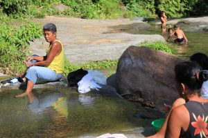 Im VG Frauen, die mit Seife ihre Wäsche in einem Bach waschen, im HG spielen Kinder im Wasser.