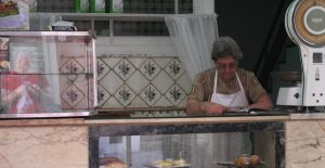 Geschäftsfrau mit Schürze hinter ihrer Backwaren-Theke in Lissabon, lesend, rechts daneben eine große Waage, im HG Fliesen