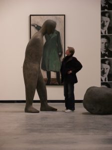 Eine Frau (im Profil) steht selbstbewusst vor einer großen Figur; dahinter Gemälde einer Person mit Hitlergruß.