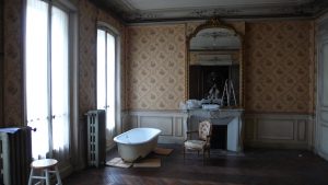 Ein unbewohntes Zimmer in Paris mit zwei bodentiefen Fenstern, großem Barockspiegel über dem Kamin; Hocker, Badewanne, barocker Sessel auf dunklem Parkett; Stuckdecke, großgeblümte Tapete über Holzvertäfelung