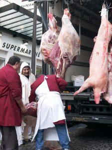 3 Metzger entladen in einer Straße in Paris große Teile von Fleisch (Schinken, Schweinehälfte) aus einem LKW. Ein Arbeiter freut sich.