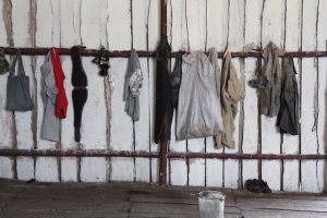 Kleidung von Holzköhlern bei Cienfuegos (Cuba) hängt aufgereiht an dunkler Holzlatte vor weißer Bretterwand, davor ein leerer Eimer.