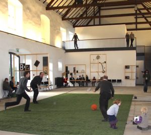 Auf einem Kunstrasen spielen 3 Männer mit einem Ball und lösen durch die Bodenkontakte Musik aus. 2 Kinder mit Ball, ringsum und auf einer Empore Zuschauer. Weitere Musikobjekte hängen im Raum.