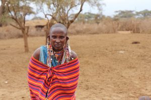 Massai-Frau mit reichem Schmuck, in buntes Tuch gehüllt (Frontalaufnahme), HG braune Erde, Bäume, Hecke