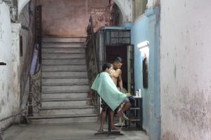 Vor einer einst herrschaftlichen Treppe karge Arbeitsstelle eines Friseurs (nackter Oberkörper) mit einem Kunden, der auf hohem Stuhl vor einem Spiegel sitzt. Beide im Profil. (Havanna, Cuba)
