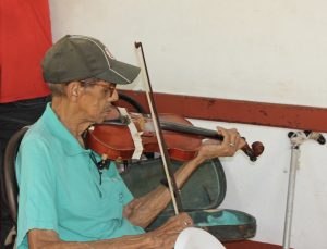 Geigenspieler (Profil, mit Schildmütze, sitzend, rechts daneben Gehstock, im HG Geigenkasten)